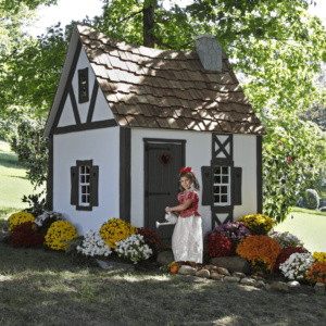 A child in princess costume stands in front of the Fairytale Cottage playhouse, surrounded by vibrant flowers and lush greenery. It's wood tile roof with faux brick chimney is reminiscent of a fairytale.
