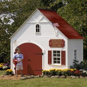 A child stands holding a toy fire helmet beside the Five Alarm Firehouse backyard playhouse, which resembles a real fire station. A Engine Company No. 8 sign adorns the white playhouse with red accents, featuring a small American flag and interactive alarm bell.