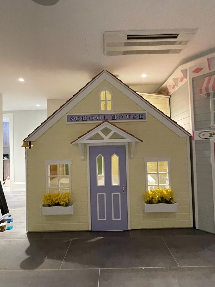 Indoor School House playhouse within a play village showcasing purple door complemented by two windows with bright yellow flower boxes.