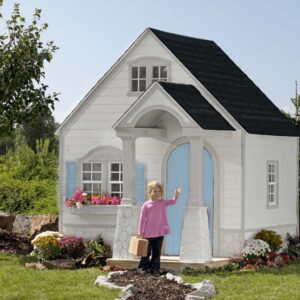 A joyful child in a pink shirt waves in front of the Storybook Bungalow, a charming white playhouse with blue shutters and door, surrounded by vibrant plants and flowers.