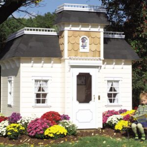 A child sits near the Victorian Mansion playhouse, styled like a mini Victorian home. Its surrounded by vibrant flowers and features a tower, white trim, and an elegant glass-paneled front door with a stained-glass accent above the door. This sweet cubie space is decorated with curtains lining the operable windows.