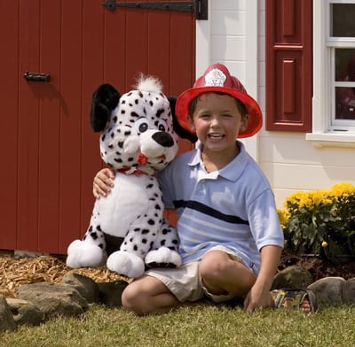 A child wearing a firefighter hat kneels beside a stuffed dalmatian in front of the backyard Five Alarm Firehouse playhouse.