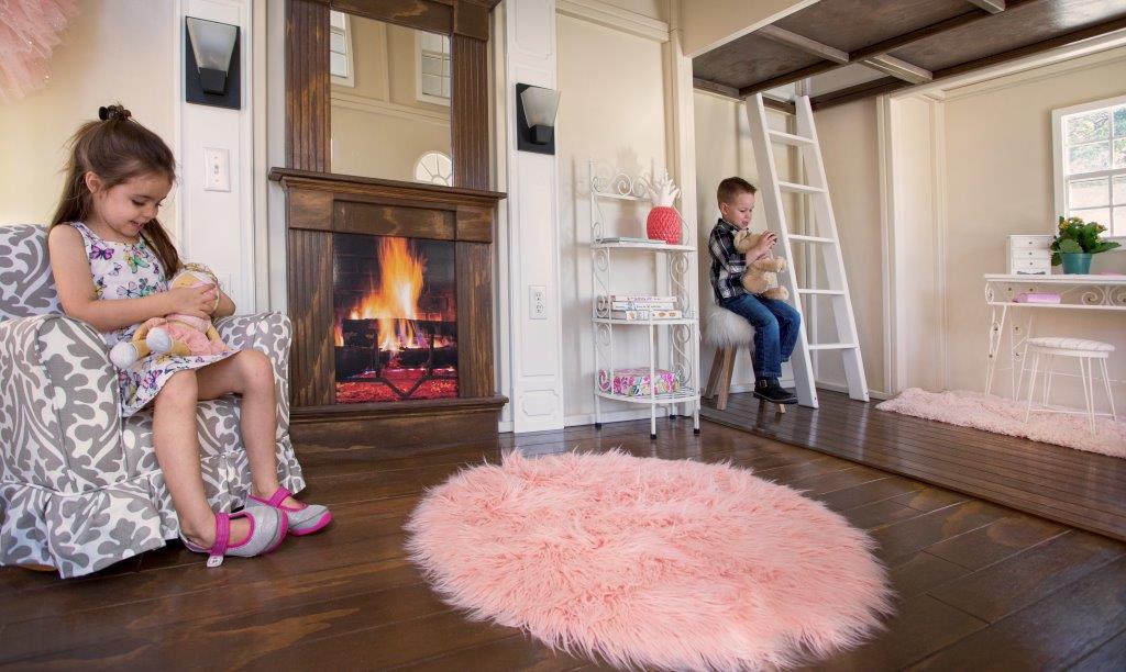 In Casa de Caramel playhouse, a girl on a chair and a boy hold stuffed animals as they enjoy the warmth of a faux fireplace. Also shown are the featured hardwood floors, and white ladder which leads to the second floor.