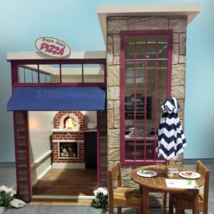 A model of Uptown Pizza Shop shows it's detailed stone facade, large windows and awning. While the inside features a brick oven with a flame feature, hand-built, child-sized wooden counters, and a menu board. A sign above the entrance reads Papa Jims Pizza.