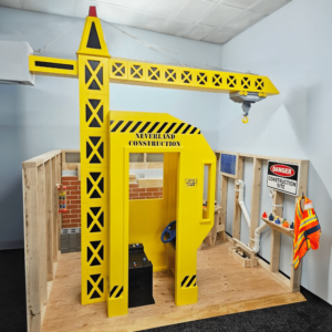 Main Street Heavy-Duty Construction Site play structure featuring bright yellow crane. Includes a wood floor, construction-themed decor, such as a warning sign, and a child's safety vest.