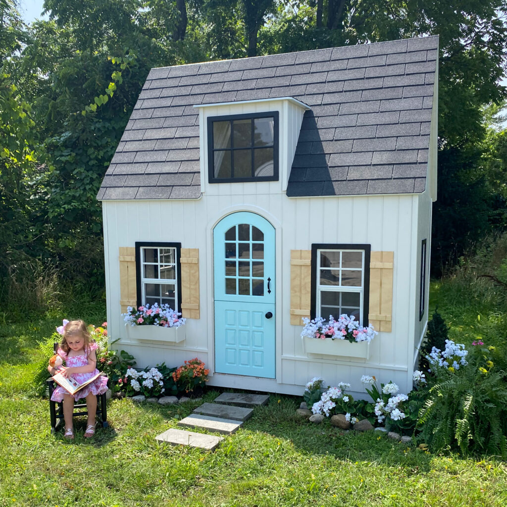 Hopscotch Hideaway with girl reading in front of the playhouse and flowers in the window boxes