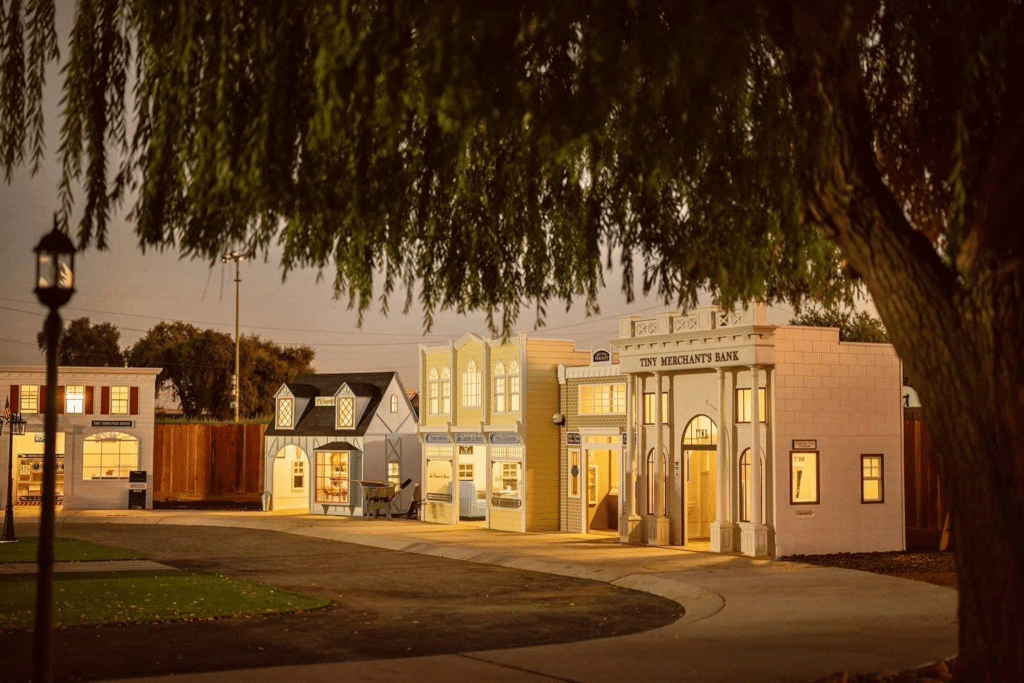 A small, illuminated outdoor play village with custom commercial playhouses designed as a bank and shops, all viewed at dusk under a large tree.