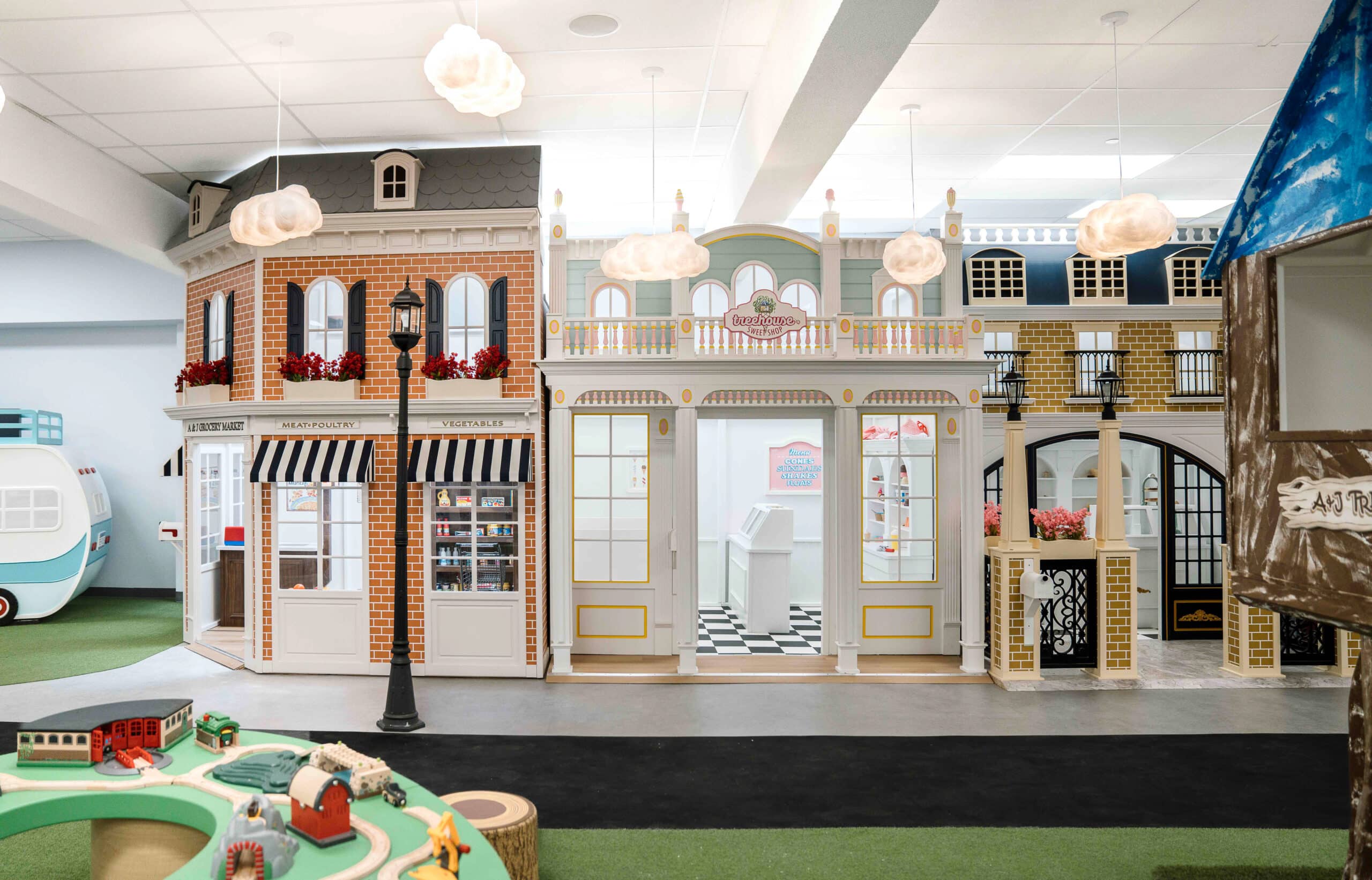 A corner grocery market playhouse with awnings and flower boxes sit next to a exquisite ice cream parlor playhouse as well as a sophisticated bistro playhouse as part of an imaginary play town in a commercial play center.