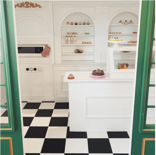 Interior of shot of the Main Street Bistro playhouse with a checkered floor, white shelves displaying various toy pastries, cupcakes, and a counter with a cake and bell. A faux oven for imaginary play can also be seen on the left.