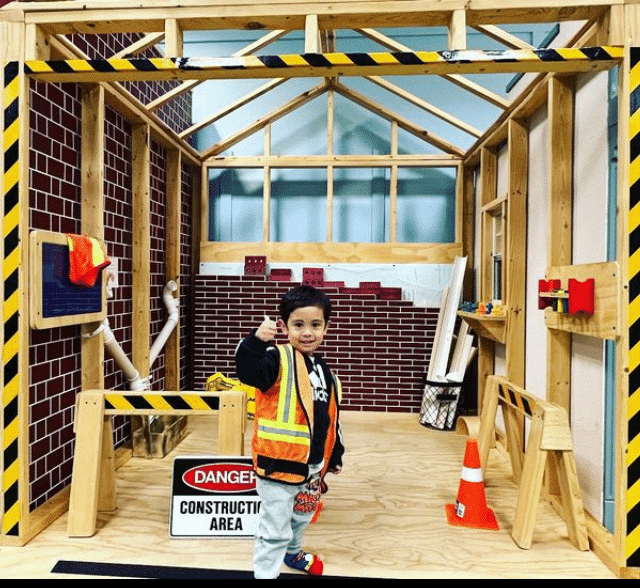 Child in a construction-themed playhouse, wearing a safety vest and holding a tool, surrounded by wooden structures, caution tape, and construction signs.