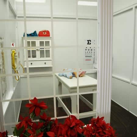 Looking through the window of a Doctor's office playhouse where an examine table, medicine cabinet, eye chart and defribulator are ready for some imaginary play with children.