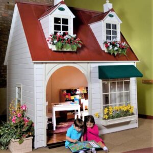 Two children sitting at the entrance of the Main Street Bookstore, looking at a book. The playhouse is white with a red roof, flower boxes, and a small interior visible through the open door.