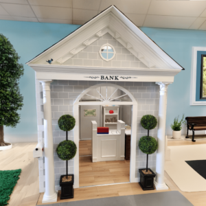 Exterior shot of the Main Street Bank playhouse with a peaked roof and columns. It features a reception area inside with a teller counter, toy cash register, and shelves fool of fake dollar bills for imaginative play. Two potted topiary plants flank the entrance.