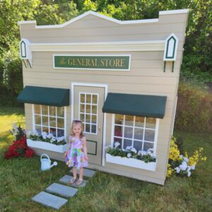 A child stands in front of an outdoor playhouse designed as a general store or market, with awnings and flower boxes.