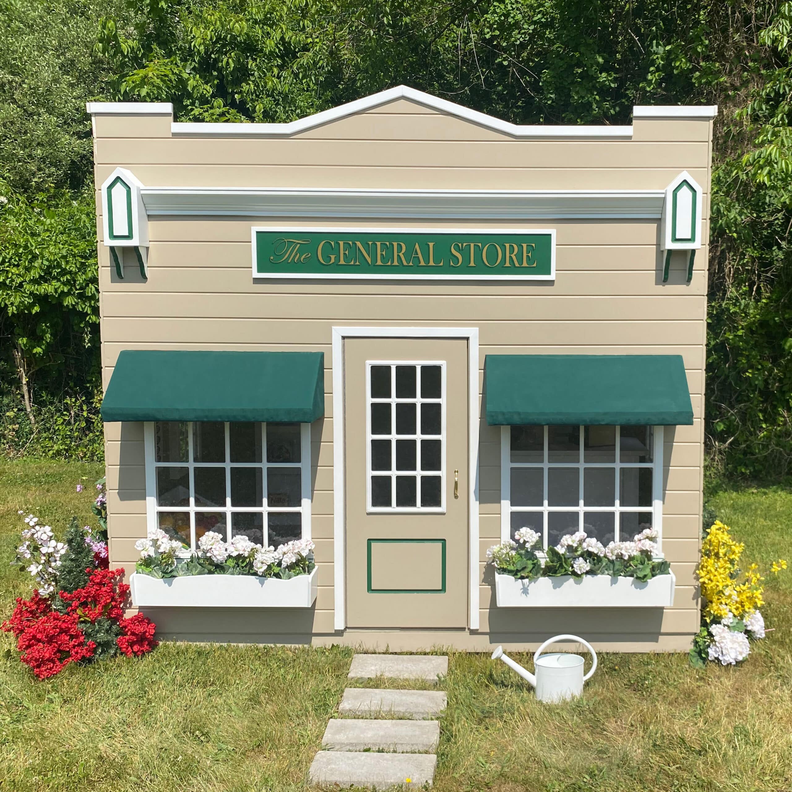Small model of a general store market with green awnings and window boxes filled with flowers, situated on a grassy backyard area with surrounding greenery. This playhouse is ready for children to come and enjoy playing "store."