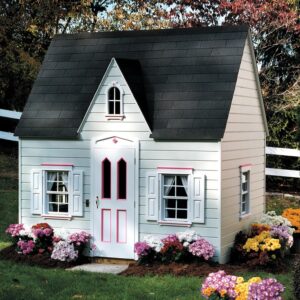 A children's outdoor playhouse, referred to as a Princess Cottage, with pink trim, featuring a steep roof and decorative shutters, is surrounded in the family backyard by colorful flowers.