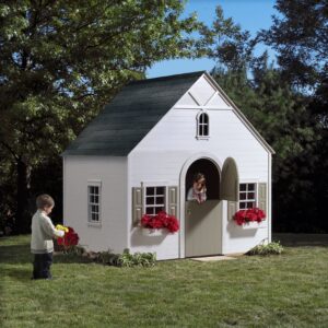 A child approaches from across the lawn as a young girl looks out the doorway of a charming cottage style backyard playhouse with shutters and colorful flower boxes.