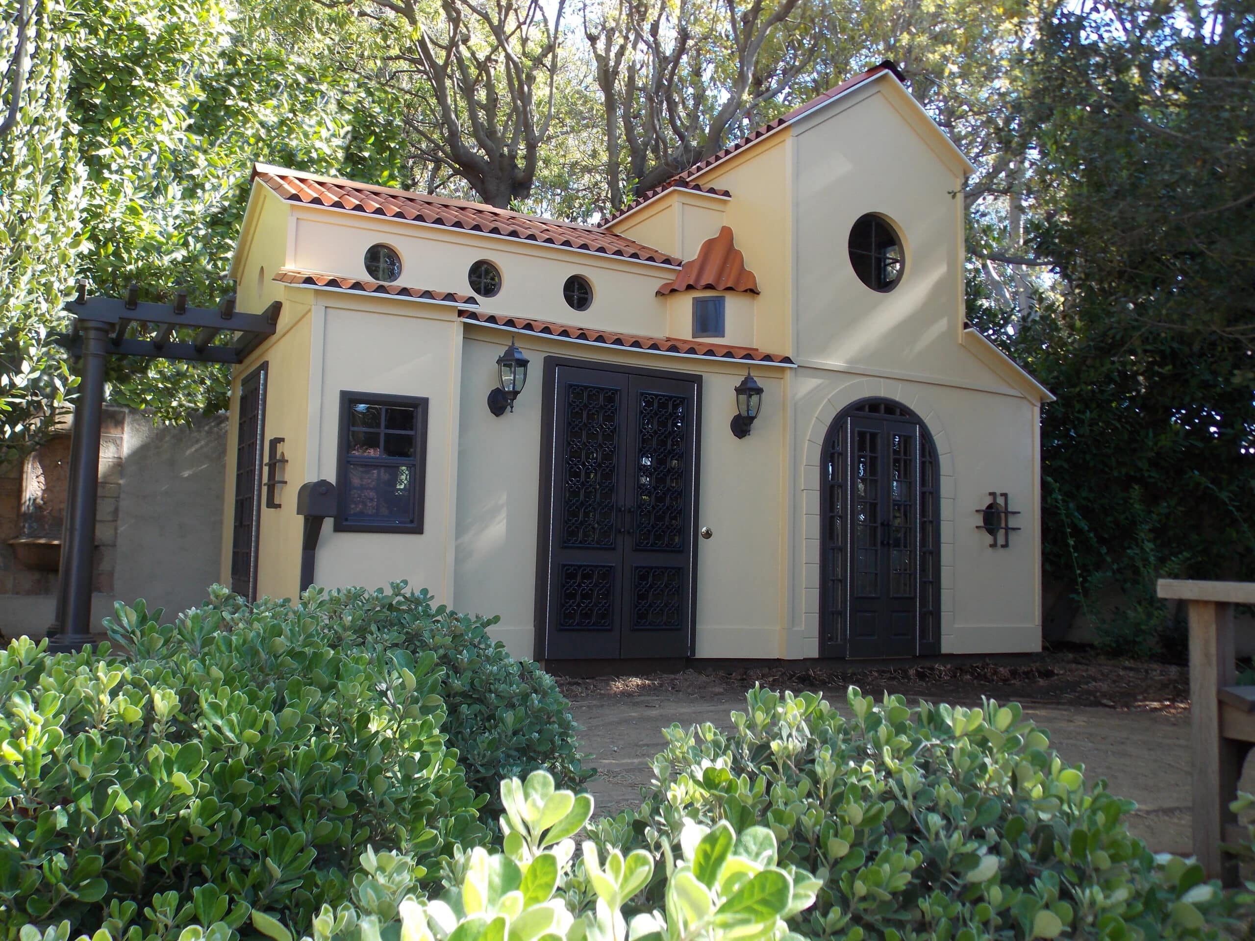 A small, light yellow custom playhouse with a red-tiled roof, surrounded by greenery. It features round windows, ornate black doors, and outdoor lanterns under a clear sky—truly a gem in the residential gallery of quaint homes.