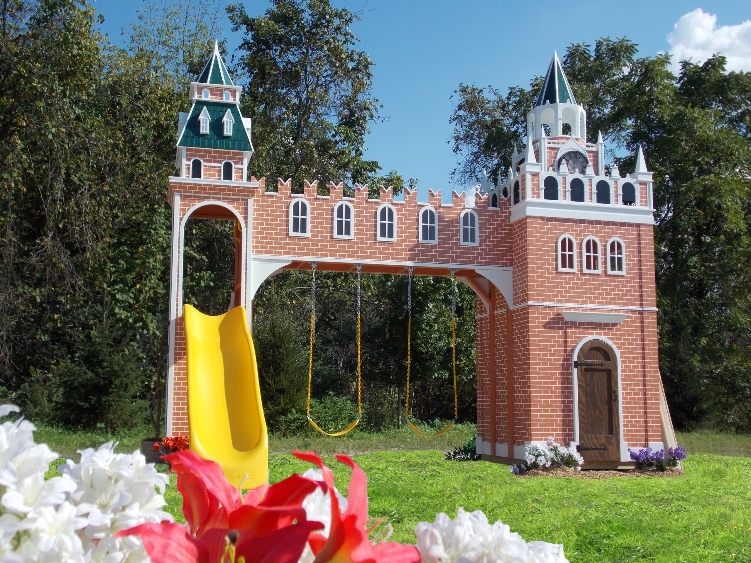 A custom playground structure resembling a brick castle, complete with a yellow slide and swings, is surrounded by grass and flowers, creating a picturesque setting reminiscent of a residential playhouse gallery.