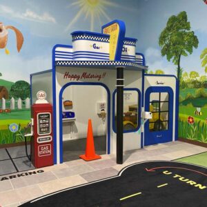 A Service Station playhouse at an indoor play facility with lovely murals behind it. It showcases retro styling, a play gas pump and a child size car inside.