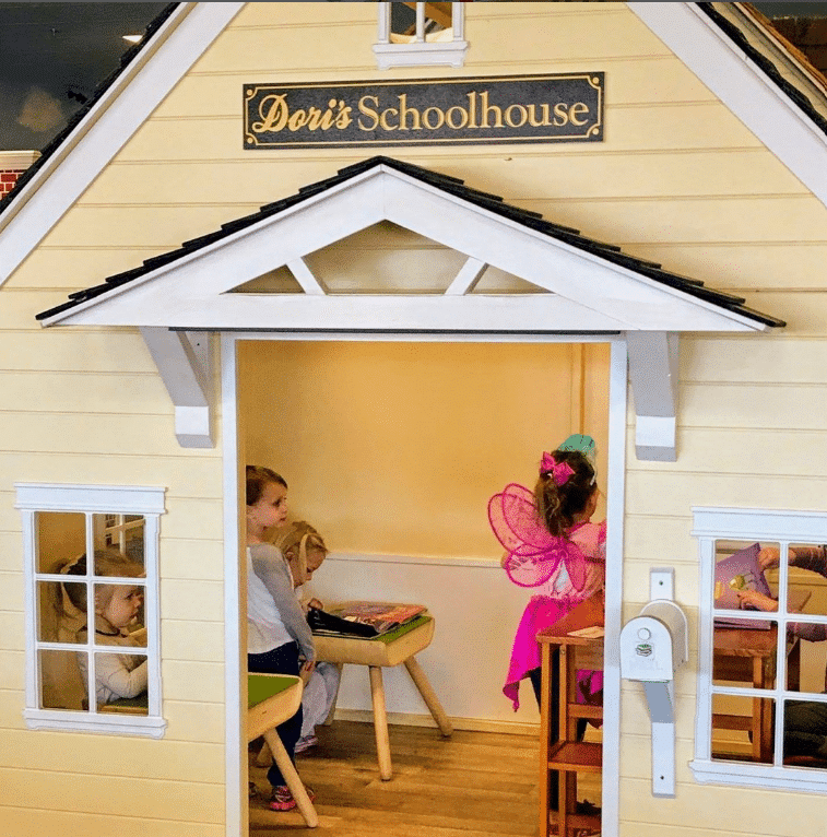 Children with little desks playing inside a schoolhouse playhouse in an indoor play facility.