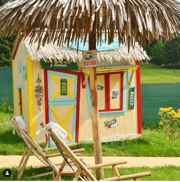 A colorful tropical style surf shack playhouse with a thatched roof, surrounded by greenery. Two wooden lounge chairs in the foreground. Signs and notices adorn the shacks exterior.