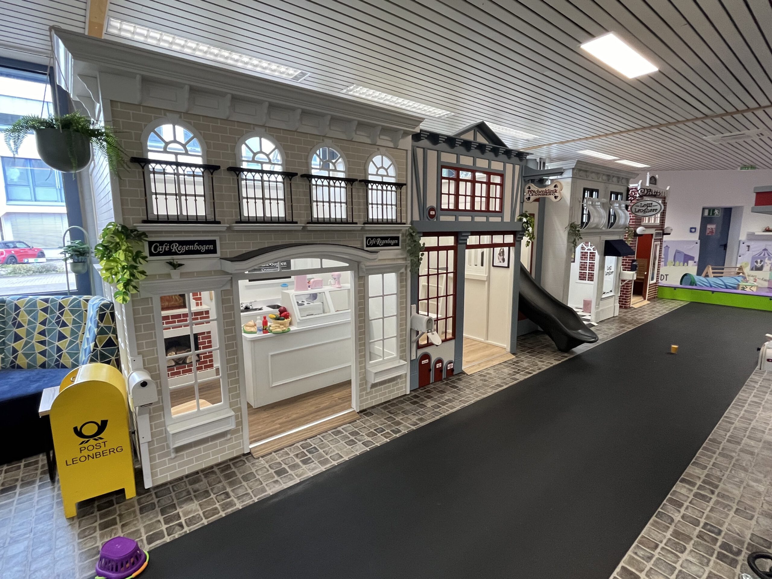 A whimsical indoor play village in Germany crafted with playhouses as a mini town. It boasts charming storefronts such as a elegantly faux brick cafe, a thrilling slide, and interactive features like a yellow postal box.