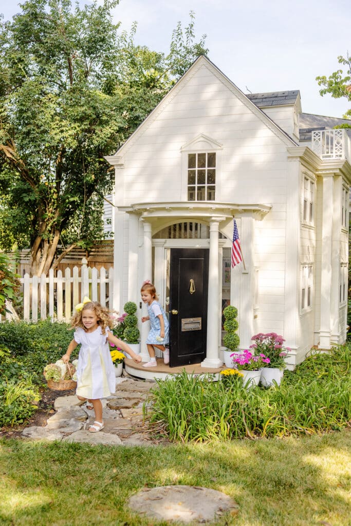 Two children walk on a stone path near a white custom luxury playhouse with columns, surrounded by greenery and flowers. An American flag hangs beside the door.
