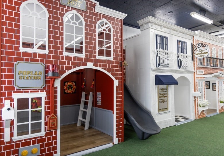 A village of imaginative play houses, inside a play cafe. The play houses include a fire station with red brick and a slide, along with a salon and cafe.