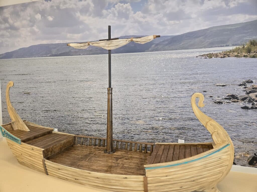 A custom built child sized boat with wood siding and a mast. There is an ocean mural behind the boat.