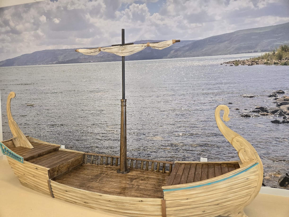 A custom built child sized boat with wood siding and a mast. There is an ocean mural behind the boat.