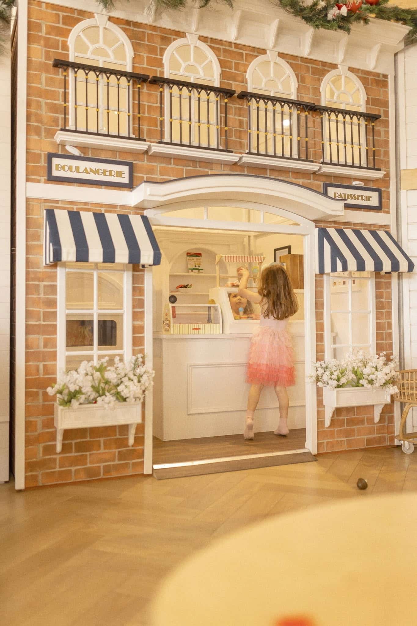 A little girl pretending to check out at a play house themed as a café, with blue and white awnings and flower boxes in the front. There are custom child sized furniture, and toys throughout the playhouse.