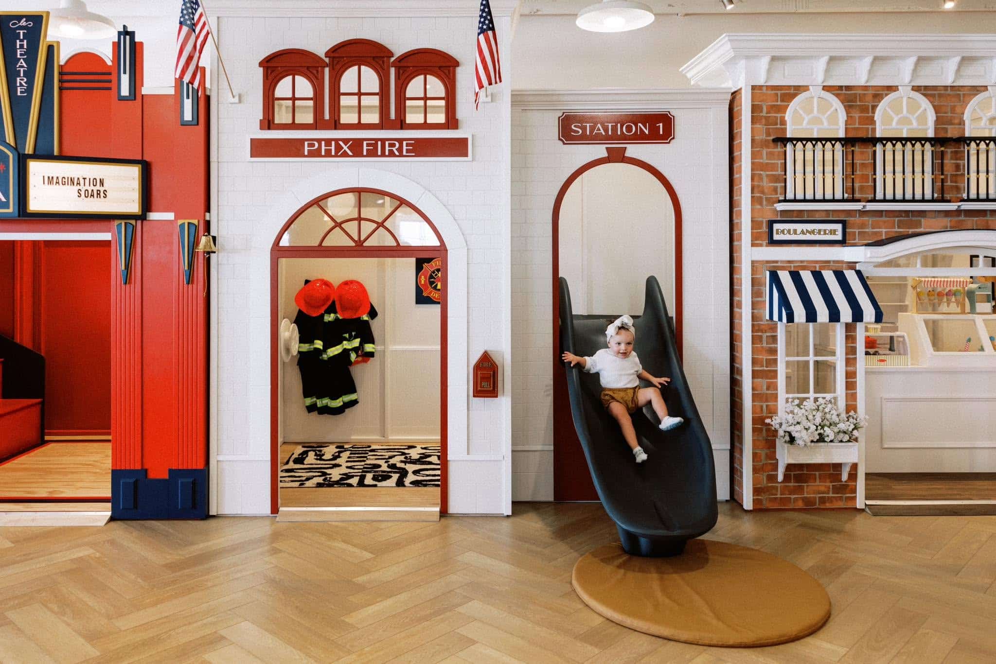 A child sliding on a slide from the wing of a fire station themed play house, inside a play café. There are other play houses on each side, including a play theater and a café.