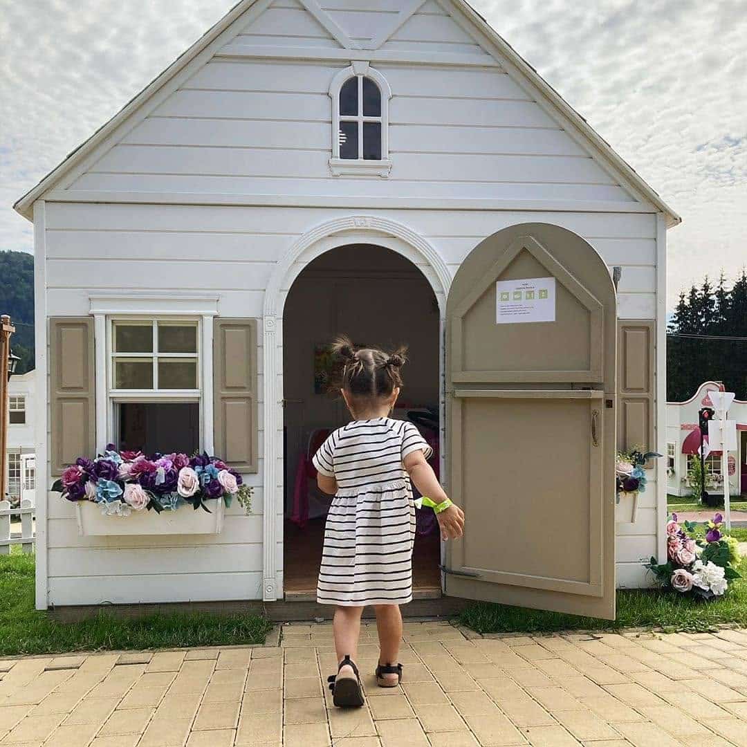 A little girl entering the front a Piccadilly play house, with a black and white stripped dress and pig tails in her hair. There are colorful flowers in the window box of the play house. It has an open Dutch door.