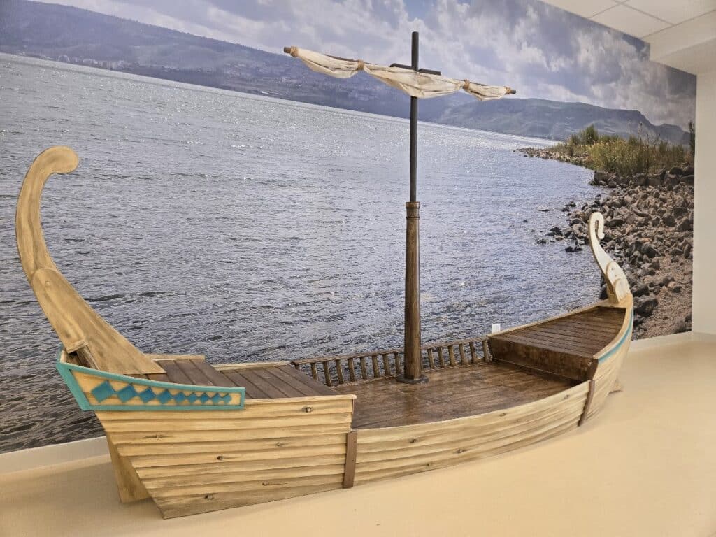 A boat, built in a miniature scale for children to enjoy, with exposed wooden sides and a rolled up sail. It sits in front of a water mural, to look like it is floating.