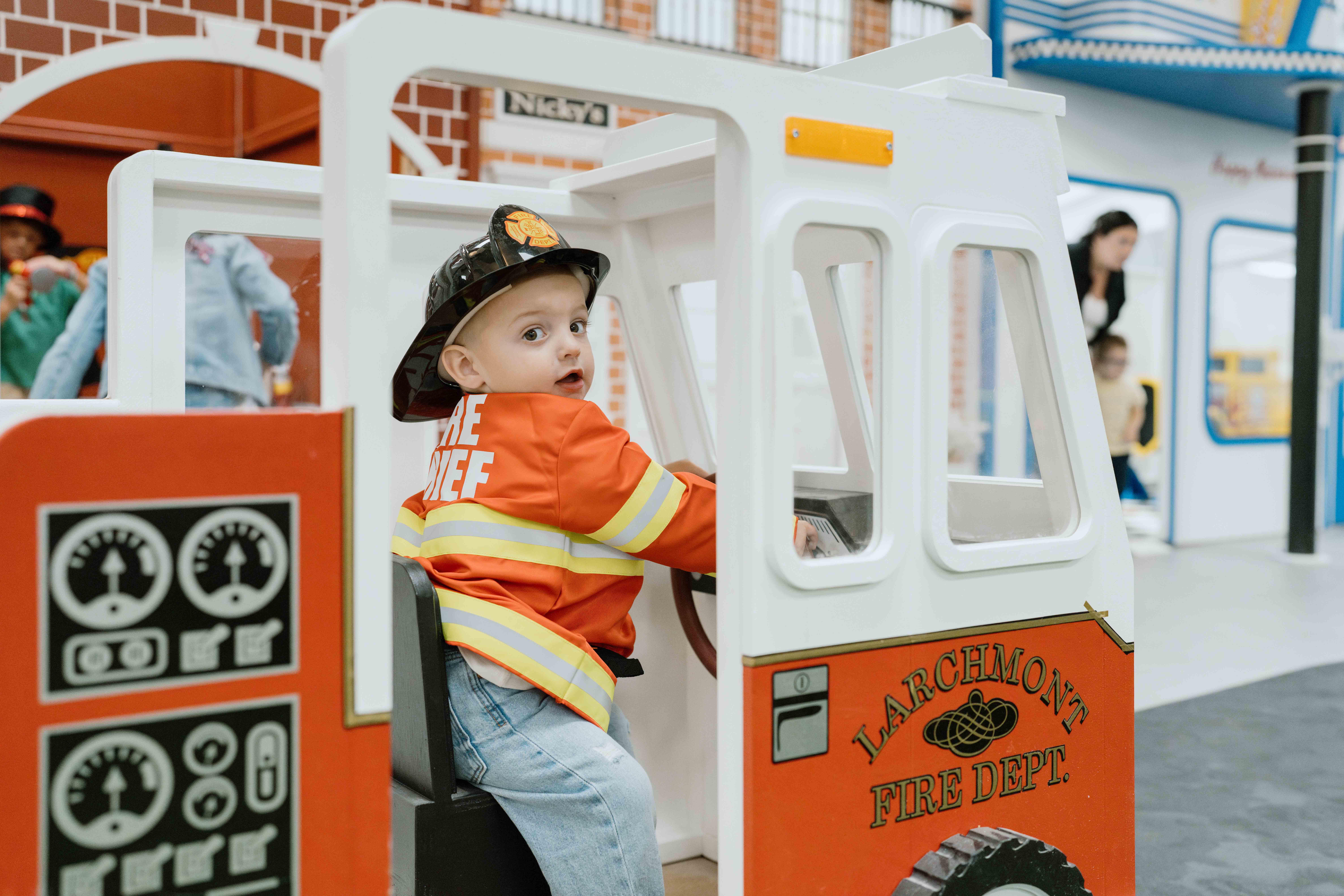 A young child wearing a fire chief jacket and helmet sits in a play fire truck labeled Larchmont Fire Dept in an indoor play area.