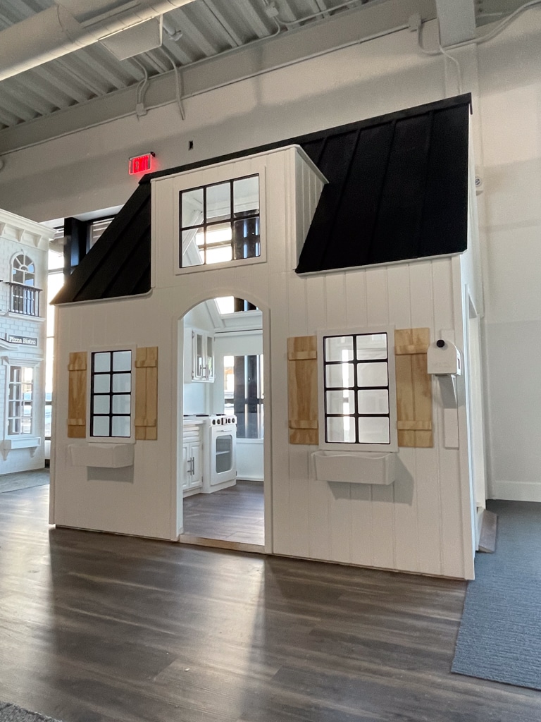 Nana's House on Main Street, a small white playhouse with a black roof, wooden shutters, and large windows, stands indoors on a wood-patterned floor with a toy kitchen visible inside.