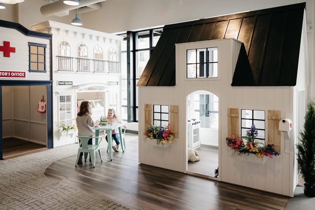 Two children sit at a small table outside of Nana's House on Main Street playhouse and other miniature buildings, such as a doctor's office and bakery.