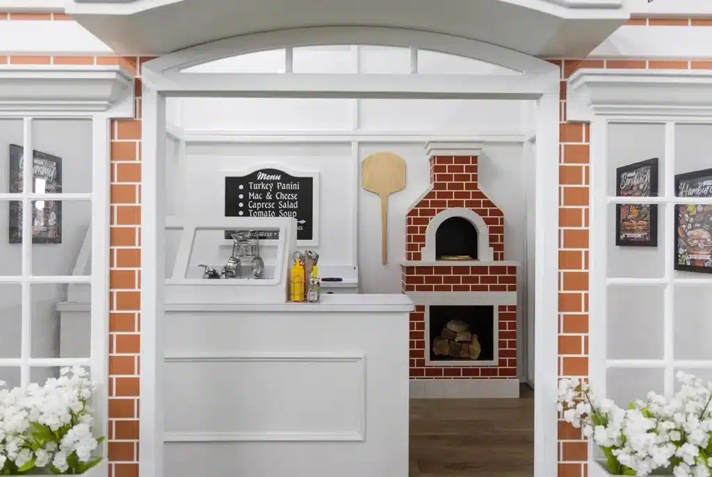 A child sized play house pizzeria setup features a front counter, a brick pizza oven with firewood, a pizza peel, and a menu board, with white flowers in planters on either side of the entrance.