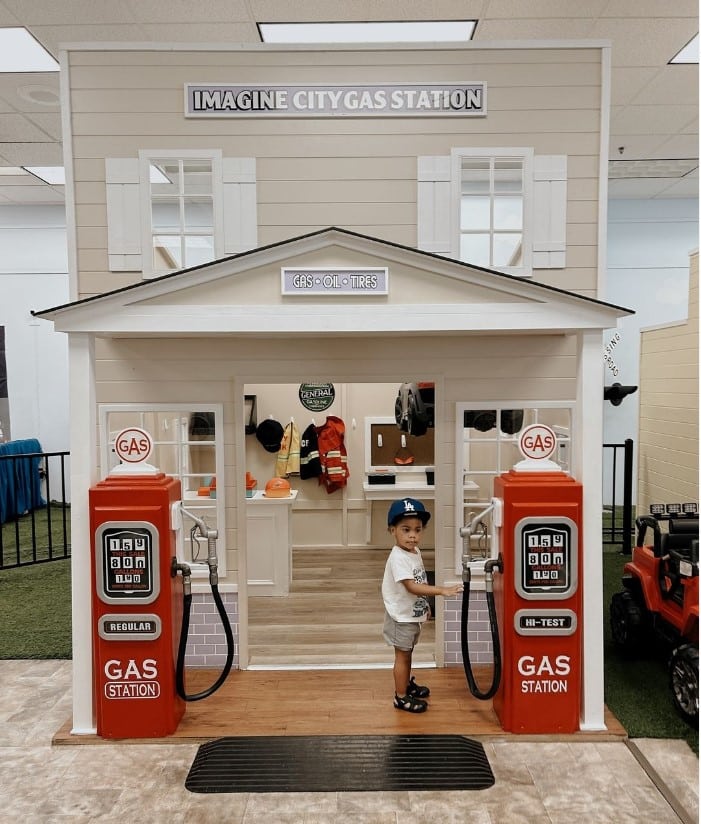 A young child stands at the Country Roads Gas Station pretend play house, holding a pump nozzle. The setup features two red gas pumps and a small building labeled IMAGINE CITY GAS STATION.