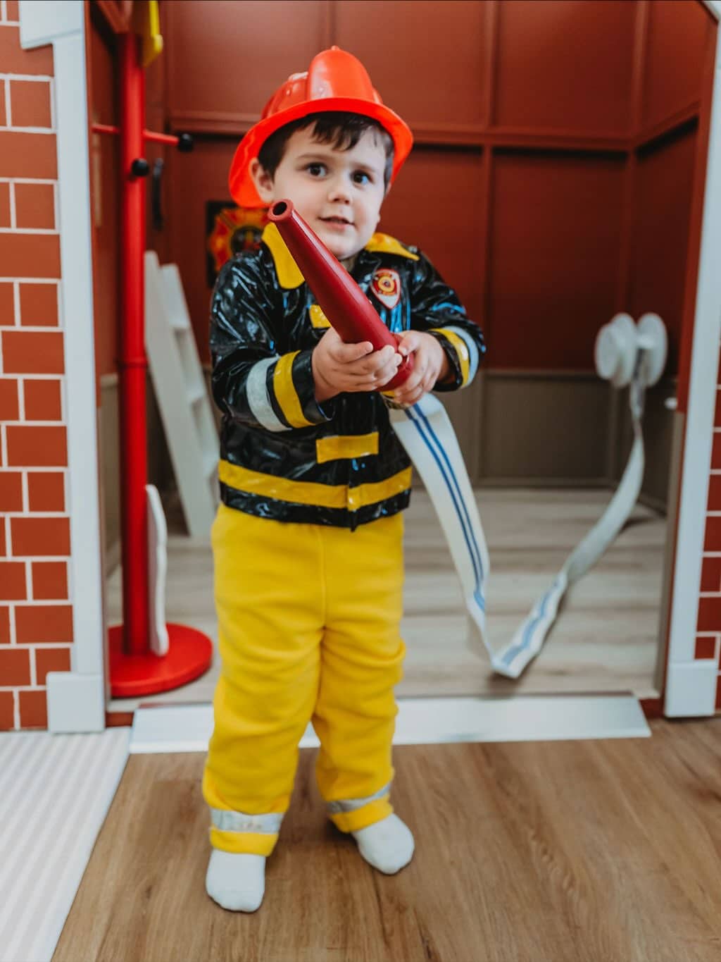 A young child dressed in a firefighter costume stands indoors holding a toy fire hose, with part of a play house themed as a fire station visible in the background.