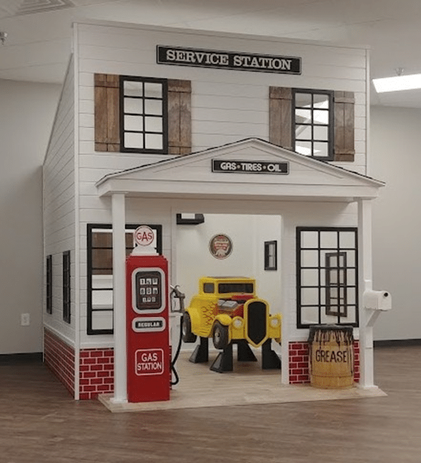 Front view of the Country Roads Gas Station, an indoor play structure for kids, designed as a vintage service station with a gas pump, yellow toy car, and signs for gas, tires, and oil.
