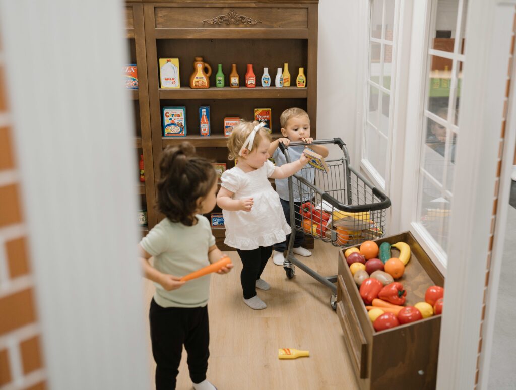 Three young children play shopping in a pretend grocery store, with toy food items and a small shopping cart.