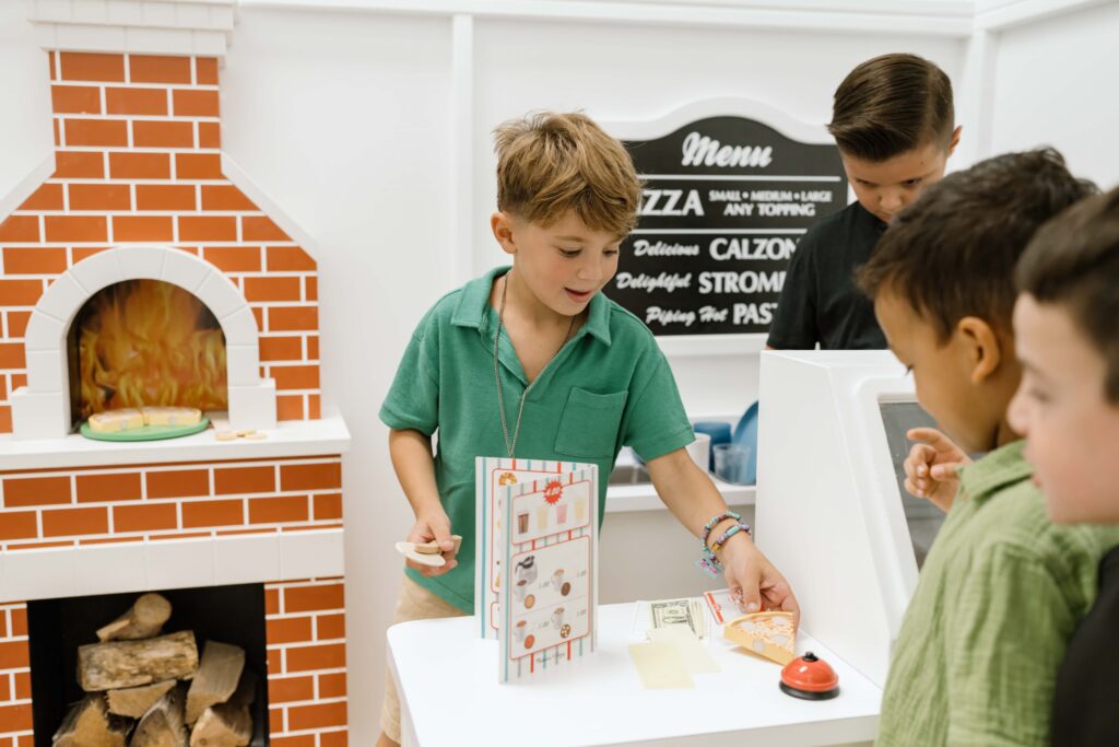 Three boys play at a pretend pizza restaurant counter with a toy oven, cash register, play money, and a pizza menu.