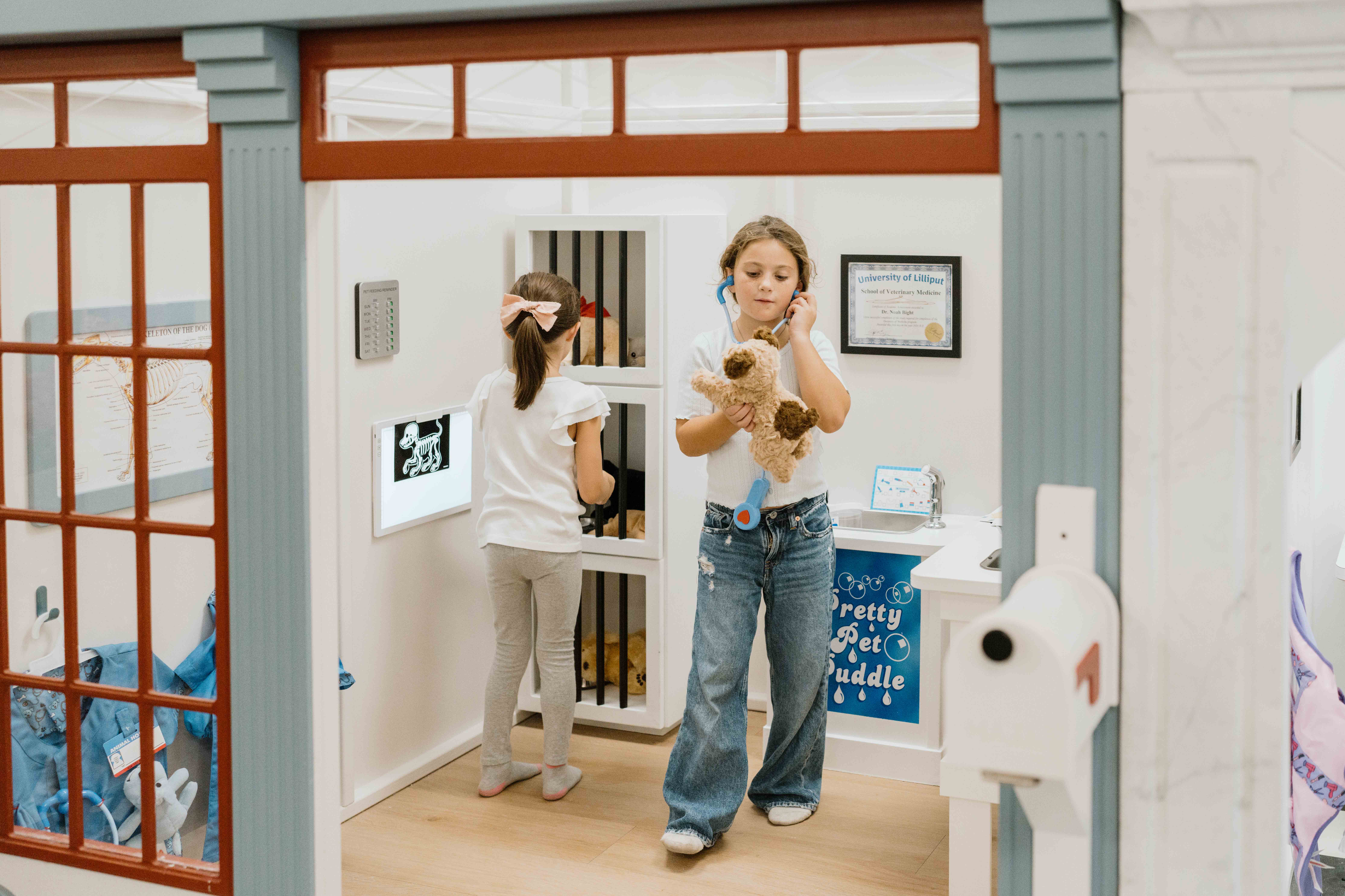 Two children play in a pretend veterinary clinic; one holds a stuffed dog, the other faces a toy animal cage. Medical toys and certificates are visible around the room.