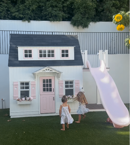 Two young children in dresses stand in front of a small white playhouse with pink accents and a slide, ready for imaginative play on the backyard’s artificial grass.
