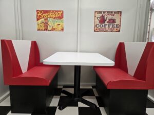 Red and white booth seats with a white table in between, set against a wall with vintage breakfast and coffee signs, on a black and white checkered floor.