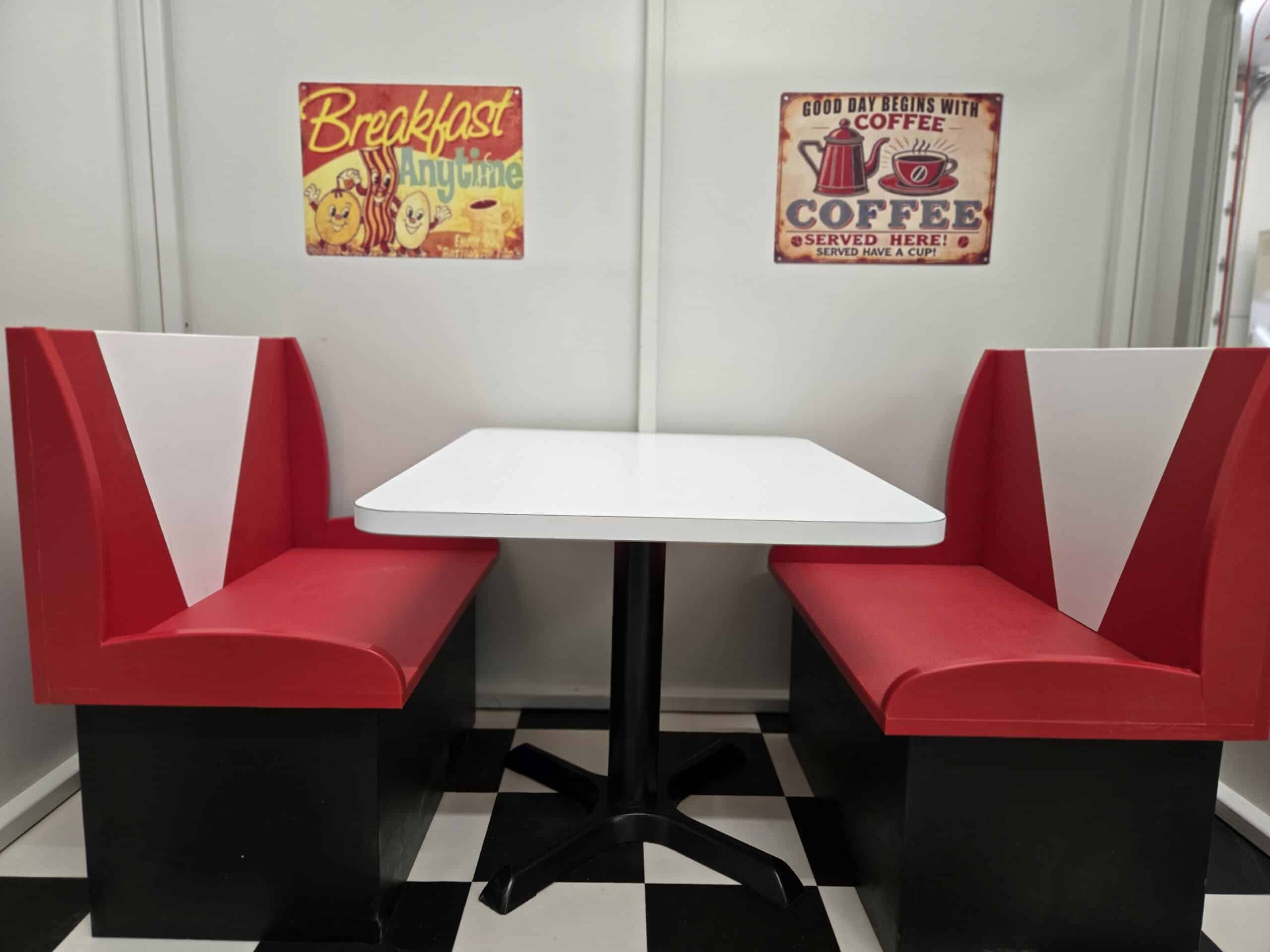 Red and white booth seats with a white table in between, set against a wall with vintage breakfast and coffee signs, on a black and white checkered floor.