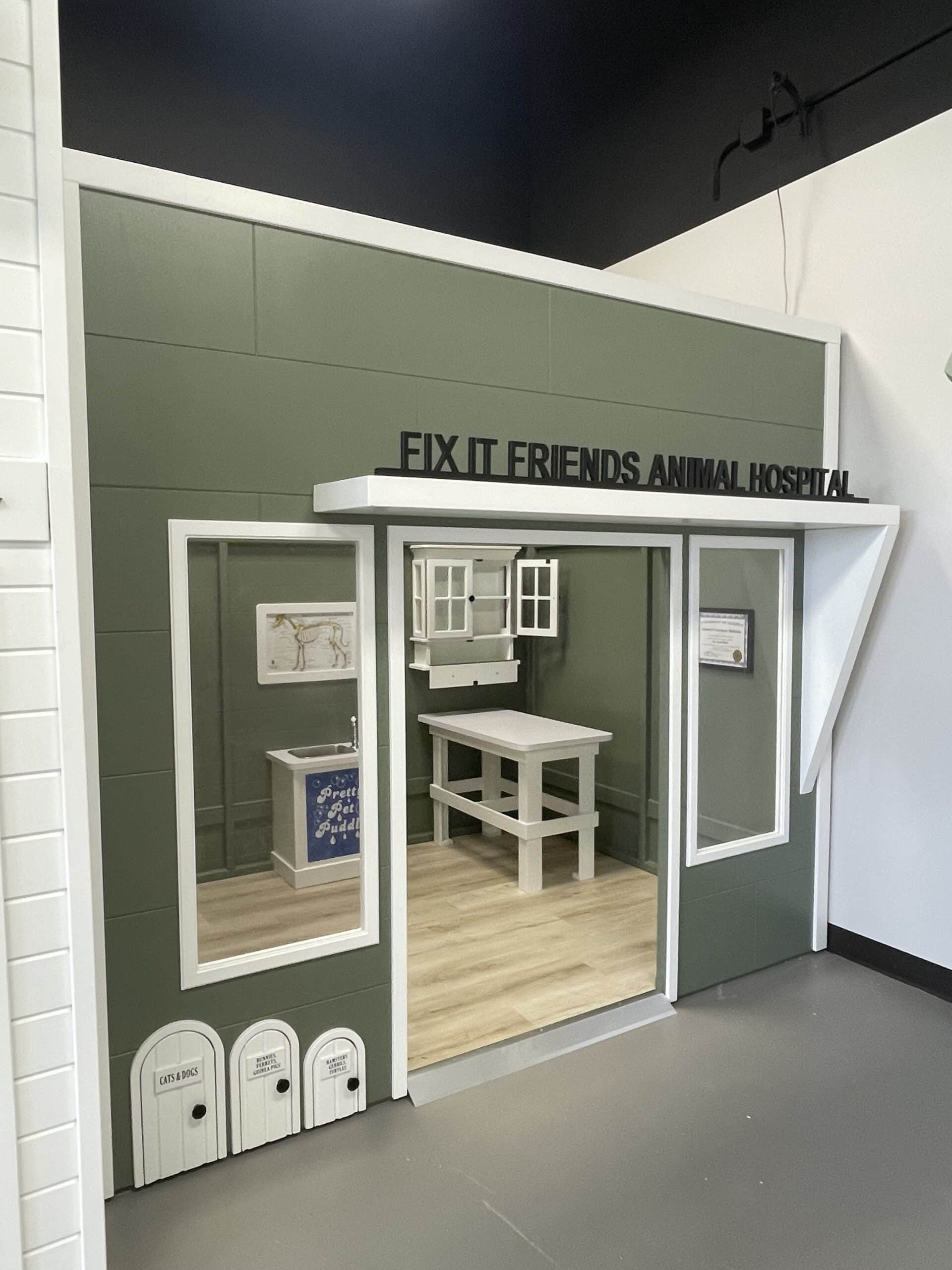 A play house themed as a veterinary clinic labeled Fix It Friends Animal Hospital with a table, counter, and pretend animal care stations inside.