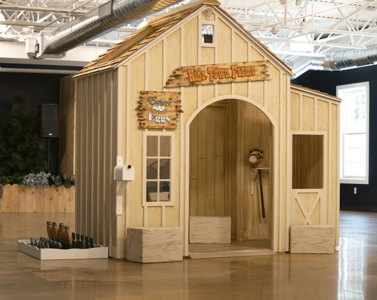 The Main Street Livery Stable playhouse is shown in an indoor play area, featuring toy vegetables, a hobby horse, and a pretend mailbox to inspire imaginative farm play.